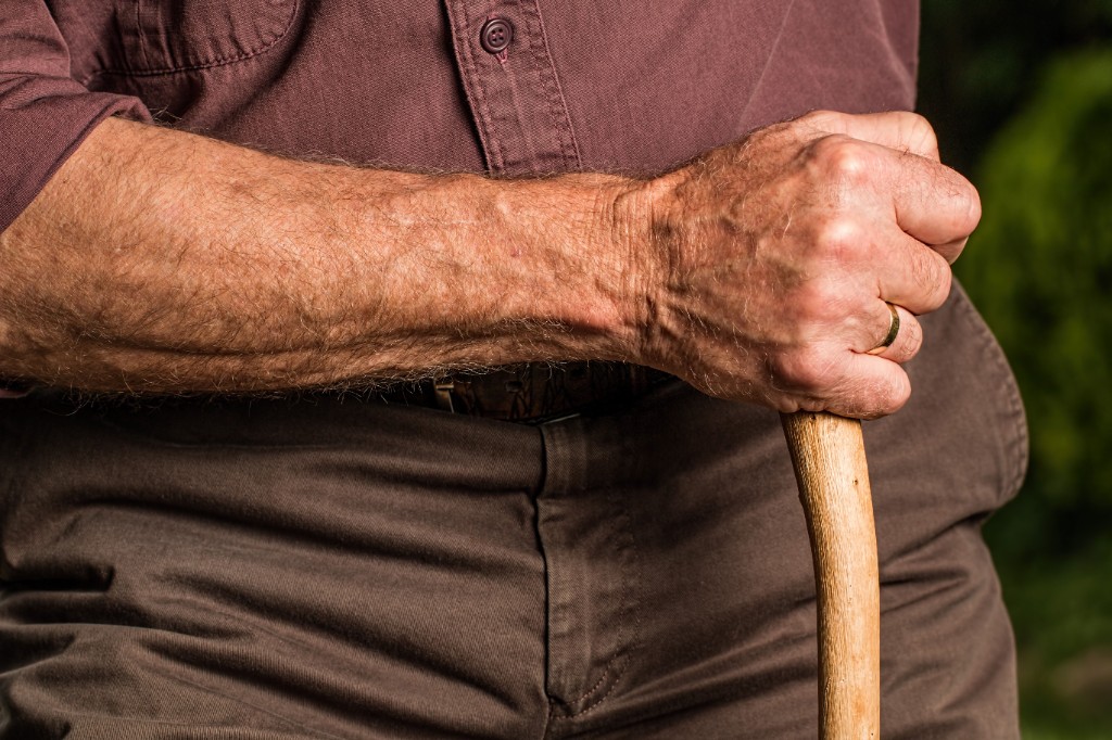An older hand resting on a walking stick — aging is visible, but independence and daily life still matter